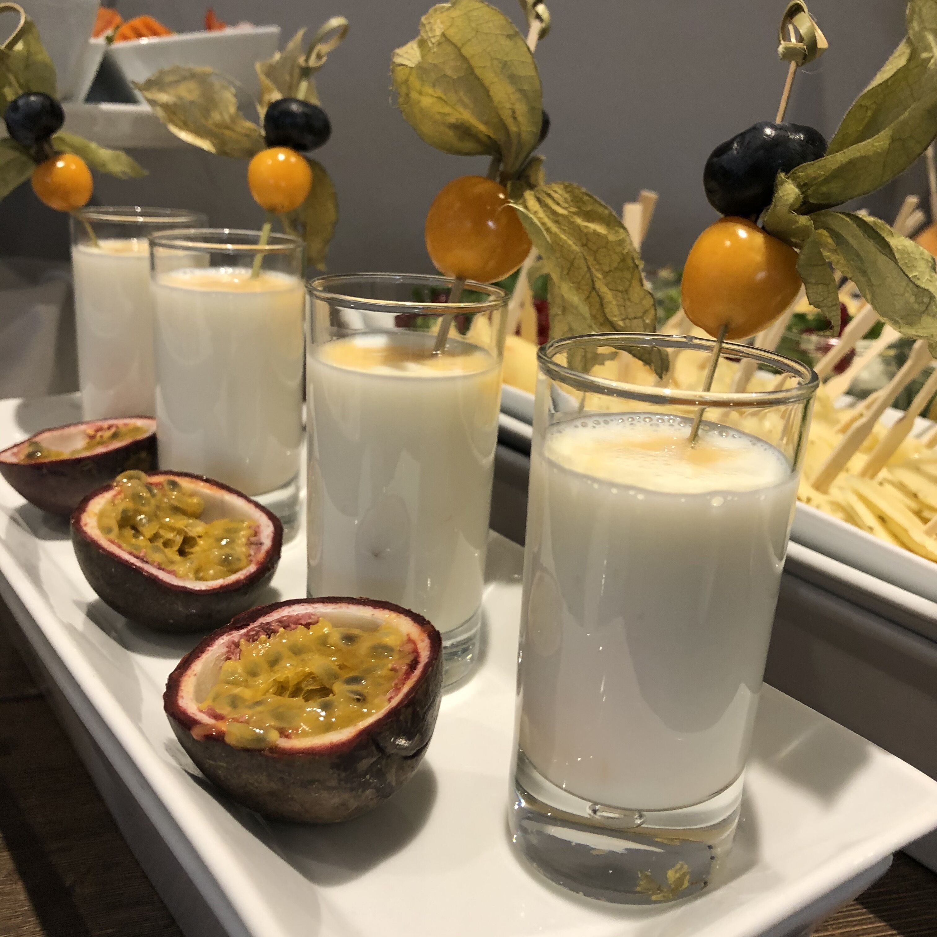 Una bandeja con vasos de una bebida cremosa, mitades de maracuyá y brochetas decorativas de frutas.
