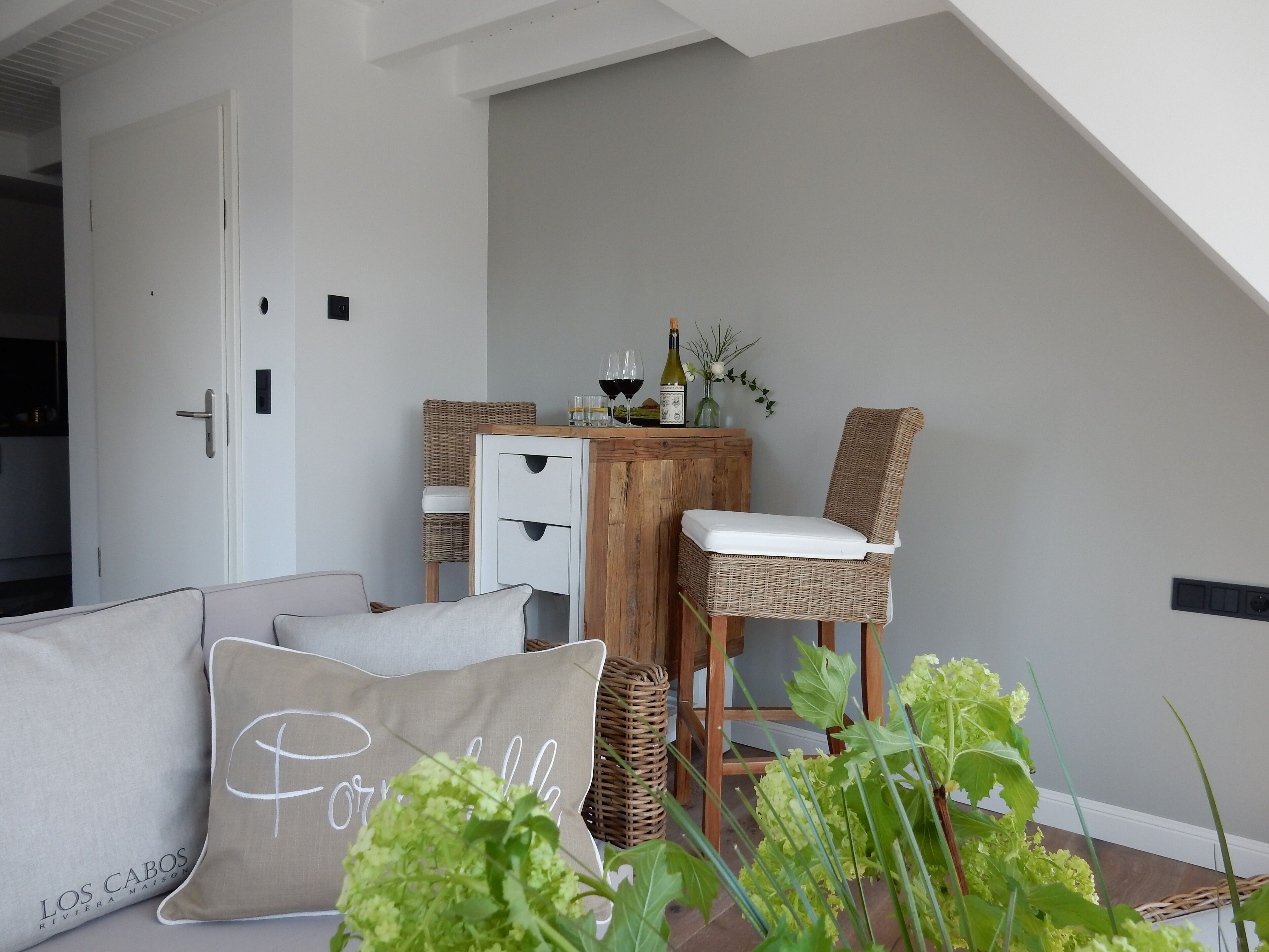A modern living room with a loft-style ceiling, a dining area with wicker chairs and a wooden table, and a cozy seating area with cushions.