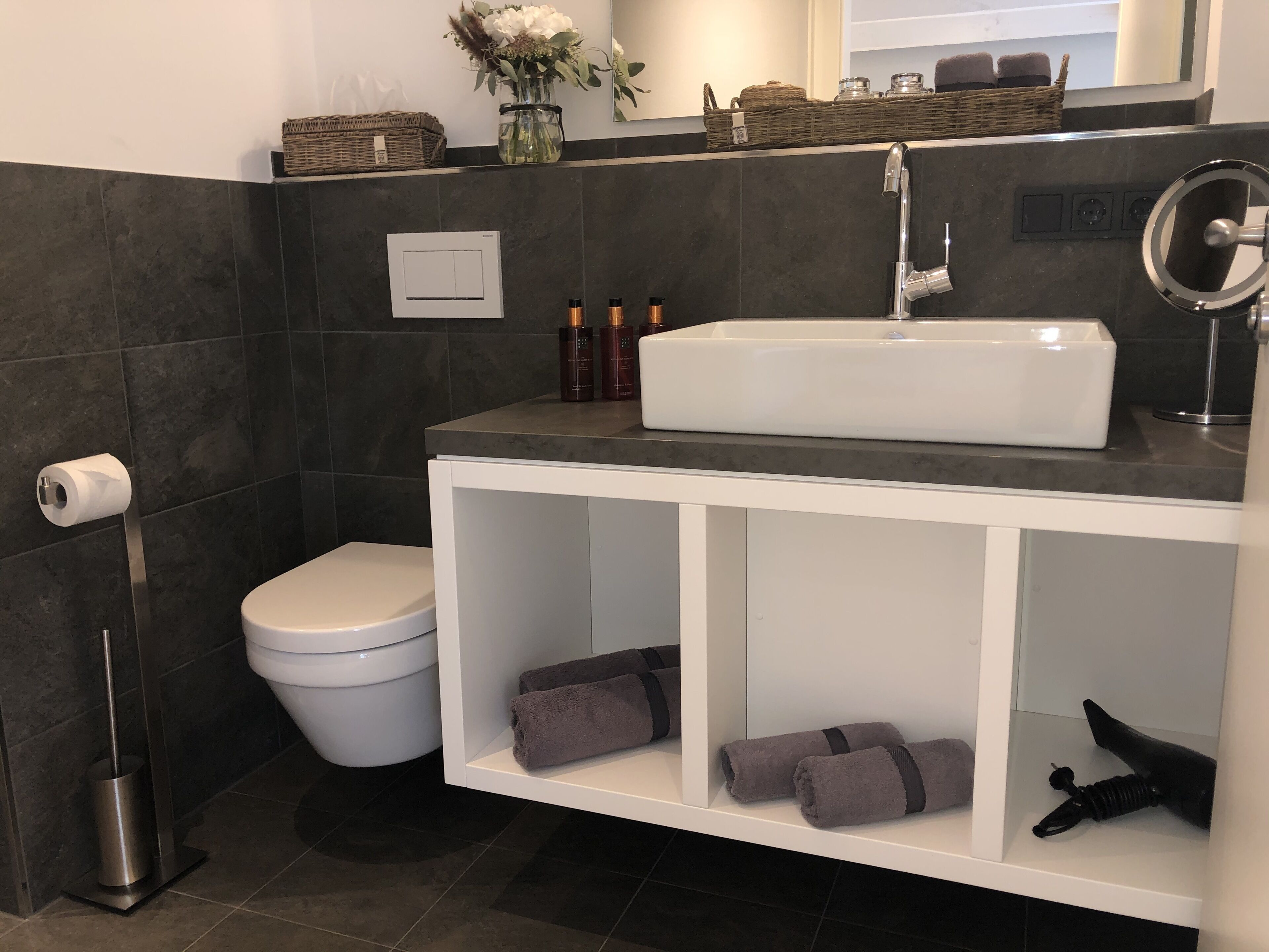 Een moderne badkamer met een witte wastafel, donkere tegels, een toilet en een plank met handdoeken.