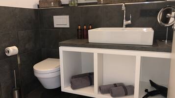 Een moderne badkamer met een witte wastafel, donkere tegels, een toilet en een plank met handdoeken.