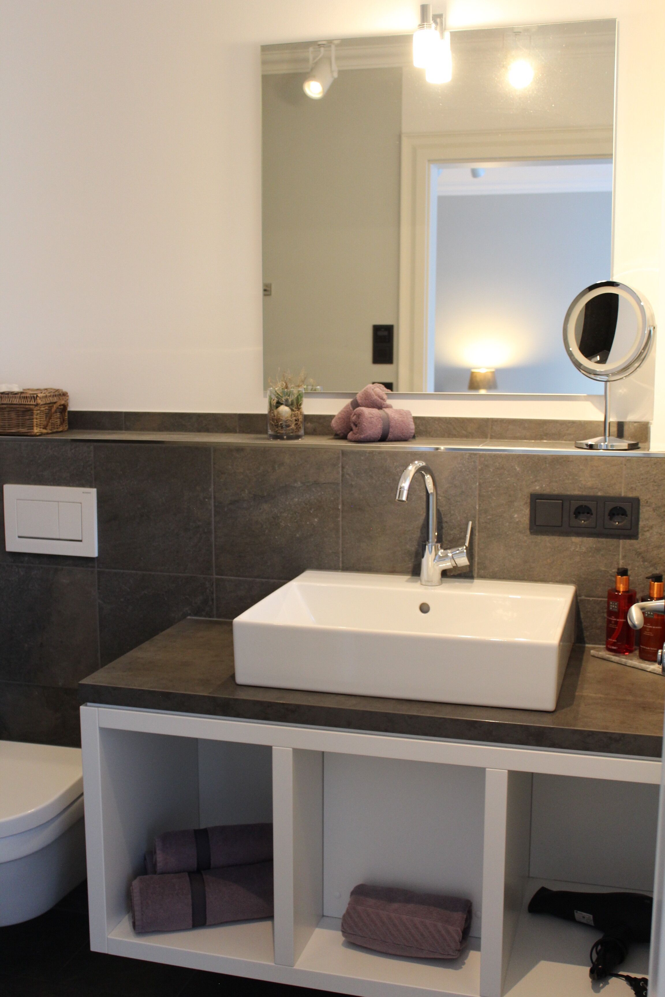 Une salle de bain avec un lavabo blanc, des carreaux foncés, un miroir et une étagère avec des serviettes.