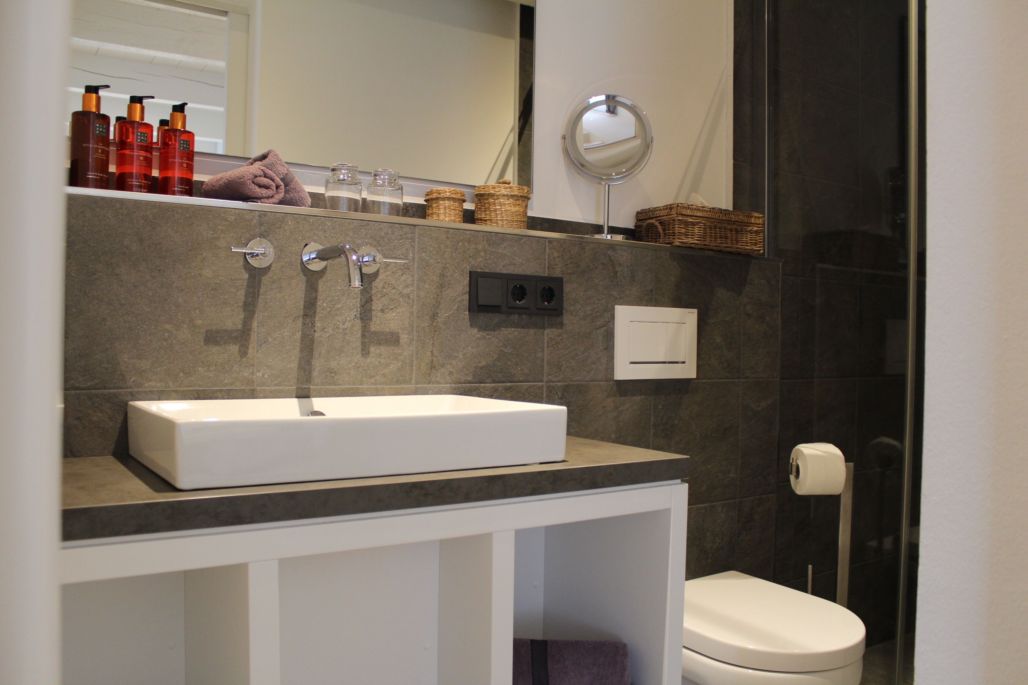 A modern bathroom with a white sink, two faucets, a mirror, and a toilet.