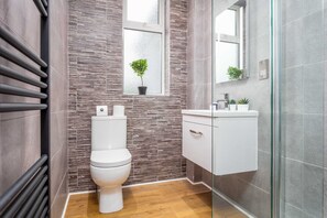 Une salle de bain moderne avec des toilettes, une petite fenêtre agrémentée d’une plante, une douche en verre et un meuble-lavabo blanc.
