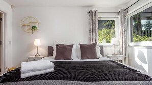 Un lit bien fait, recouvert d’une couverture noire et grise, avec deux oreillers blancs et deux oreillers marron. Une petite table de chevet avec une lampe se trouve de chaque côté du lit. La pièce a des murs blancs et une fenêtre avec des rideaux.