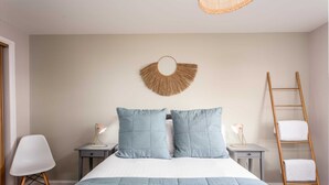 Une chambre à coucher avec un lit, deux tables de chevet avec des lampes, un objet décoratif fixé au mur, une chaise blanche et un escabeau en bois.