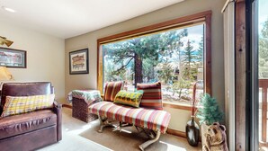 A cozy living room with a leather sofa, a striped bench, and a large window offering a view of trees and buildings. - Remodeled Cabin, Balcony. Near Walking Trails. Pool & Tennis Courts On-site (Mammoth Lakes)