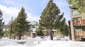 A snow-covered landscape with a building featuring a stone facade and wooden balcony, surrounded by tall pine trees and a mountain in the background. - Remodeled Cabin, Balcony. Near Walking Trails. Pool & Tennis Courts On-site (Mammoth Lakes)