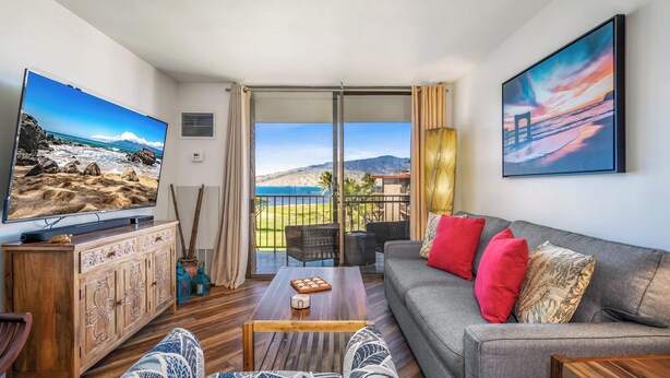 A living room with a TV, sofa, and coffee table, a balcony with a view of the beach and mountains, and a painting on the wall. - Oceanfront Penthouse Vistas (Kihei)