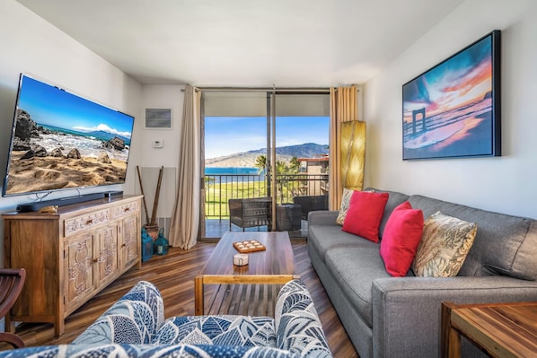 Un salon avec un téléviseur, un canapé et une table basse, un balcon avec vue sur la plage et les montagnes, et un tableau accroché au mur.