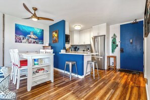 Una cocina moderna con armarios blancos, una pared de acento azul, un ventilador de techo y una zona de comedor con sillas y taburetes blancos.