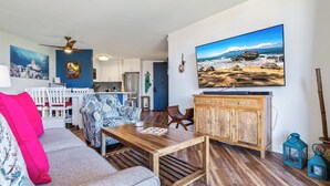 A living room with a wooden coffee table, a sofa with patterned cushions, a television mounted on the wall, and a kitchen area in the background. - Oceanfront Penthouse Vistas (Kihei)