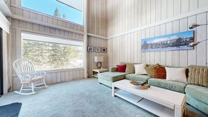 Ein gemütliches Wohnzimmer mit einem grünen Sofa, einem weißen Couchtisch und einem Schaukelstuhl. Ein großes Fenster bietet Blick auf Bäume, und an der Wand hängt ein Gemälde von einem Berg.