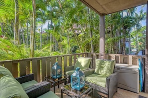A covered outdoor seating area with wicker furniture, cushions, and lanterns, surrounded by lush greenery.