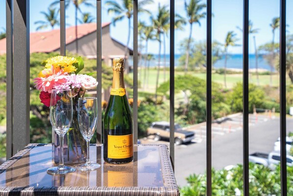 Una botella de Veuve Clicquot Brut champagne, dos copas de champagne y un florero con flores sobre una mesa de balcón con vista a un estacionamiento y palmeras.