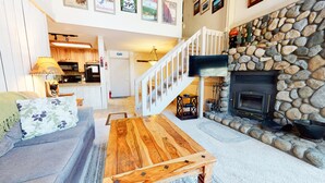 A living room with a stone fireplace, a wooden coffee table, a sofa, and a kitchen in the background.