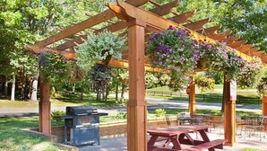Una pérgola de madera con cestas de flores colgantes, mesas de picnic y una parrilla en un parque.