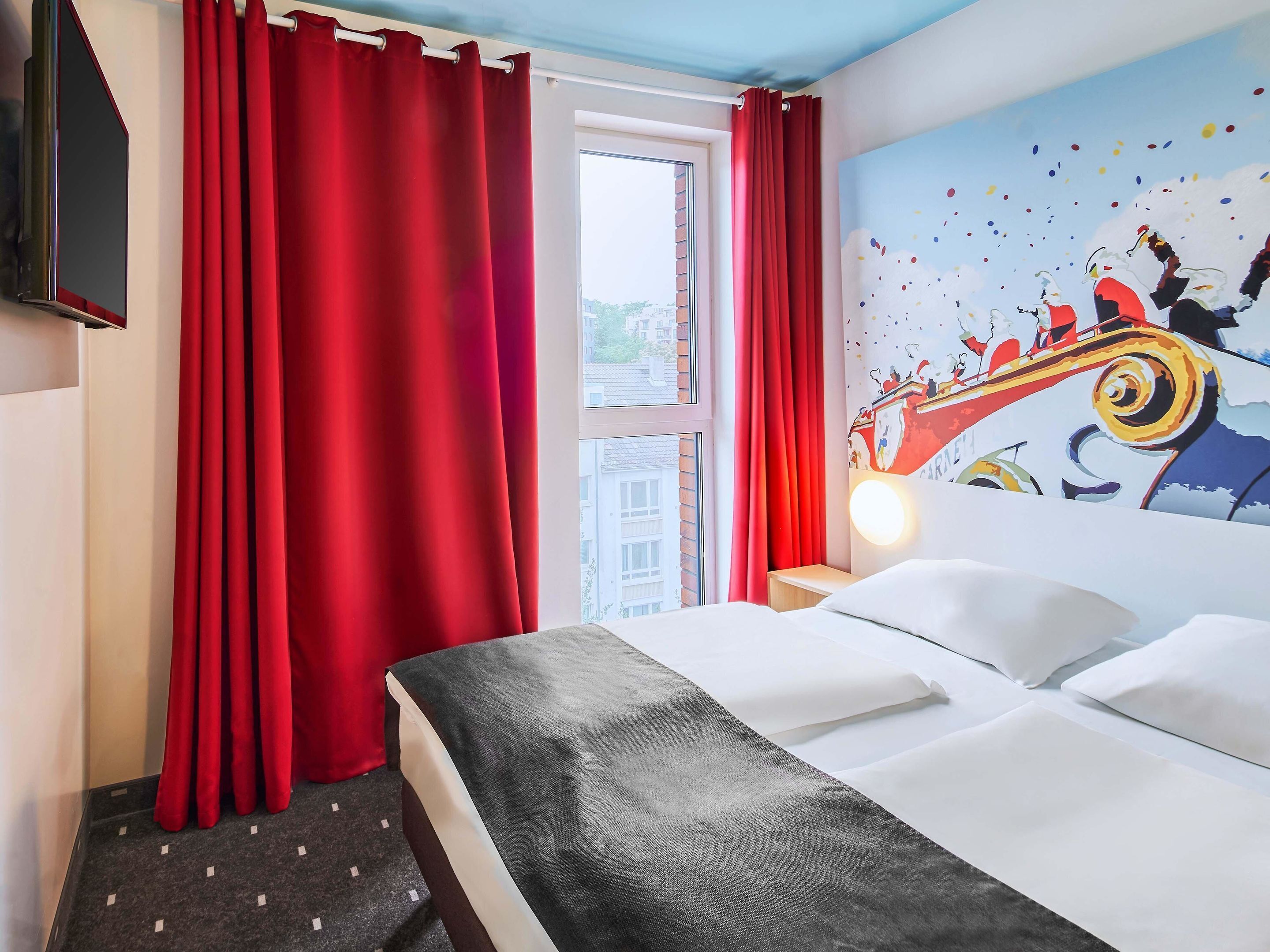 Une chambre d’hôtel avec un lit, des rideaux rouges, un téléviseur fixé au mur et une fenêtre donnant sur des immeubles.