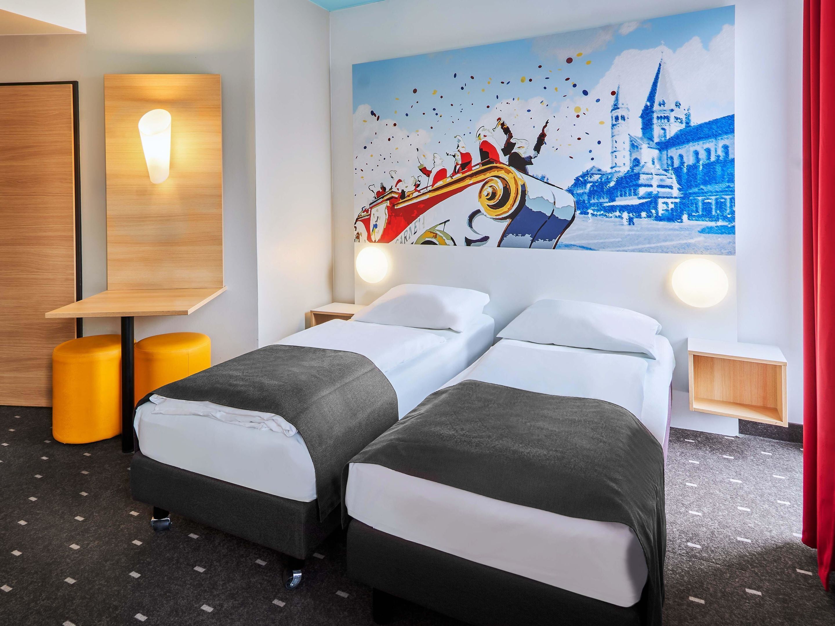 Habitación de hotel con dos camas, un escritorio de madera, un taburete amarillo y un mural en la pared.