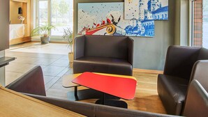 A modern lobby with leather seating, a red and yellow table, and artwork depicting a cityscape and a boat. - B&B Hotel Mainz-Hbf (Mainz)