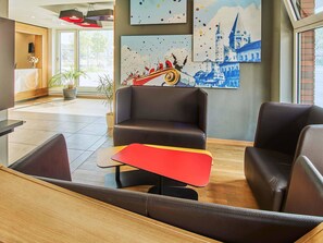 Un hall d’entrée moderne avec des sièges en cuir, une table rouge et jaune, et des œuvres d’art représentant un paysage urbain et un bateau.