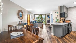 A modern kitchen with a central island, dining area with wooden chairs, and a view of the outdoors. - Refined Oceanfront Vistas (Kihei)