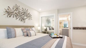 Habitación de hotel con una cama grande, un adorno en la pared, una puerta corredera que da al baño y una zona de cocina visible.