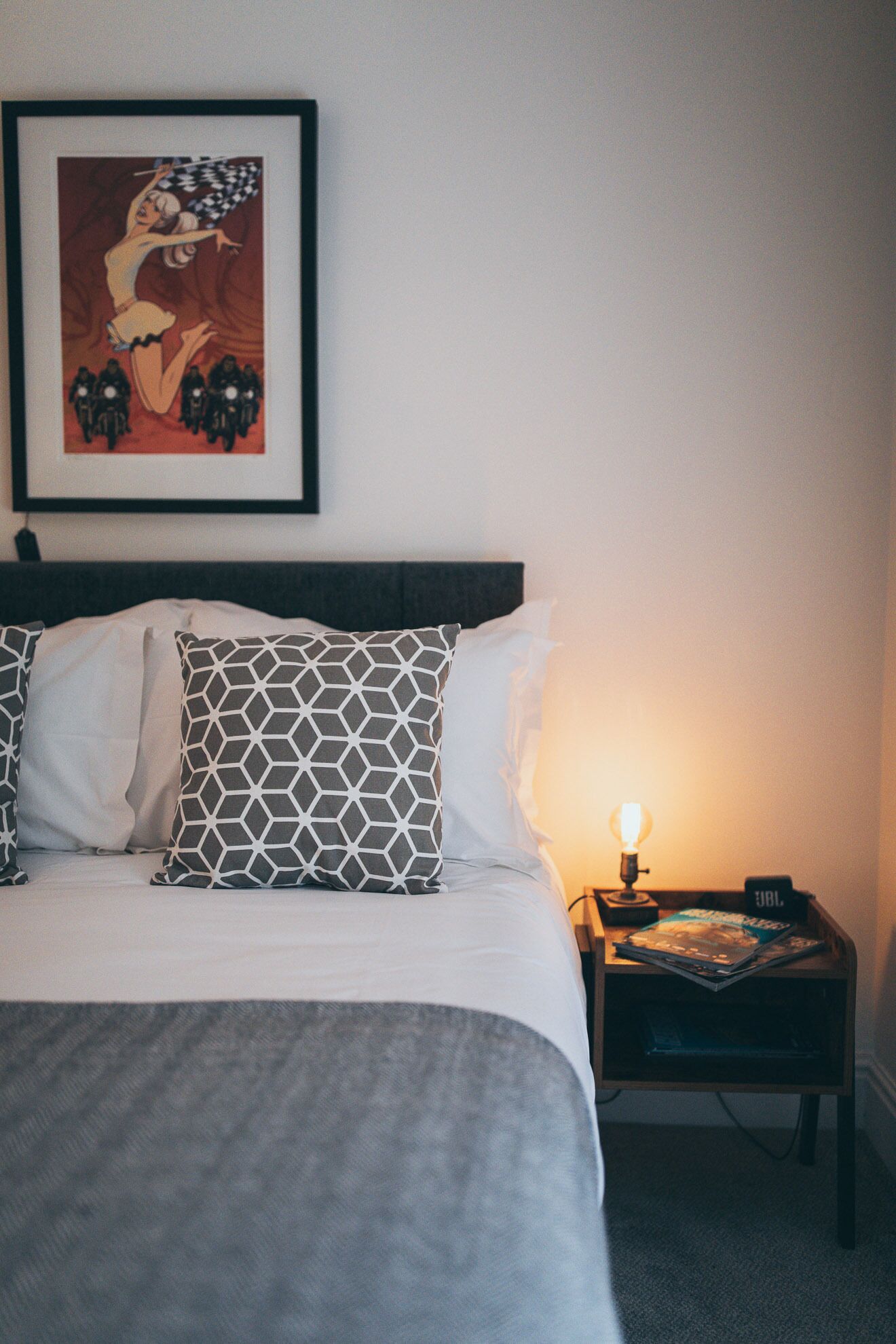 A neatly made bed with a grey blanket and patterned pillows, a framed picture on the wall, and a bedside table with a lamp and a small clock.