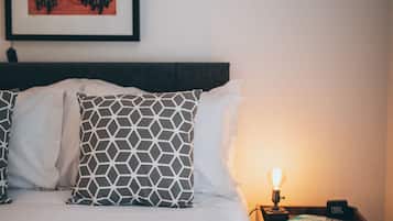 A neatly made bed with a grey blanket and patterned pillows, a framed picture on the wall, and a bedside table with a lamp and a small clock.