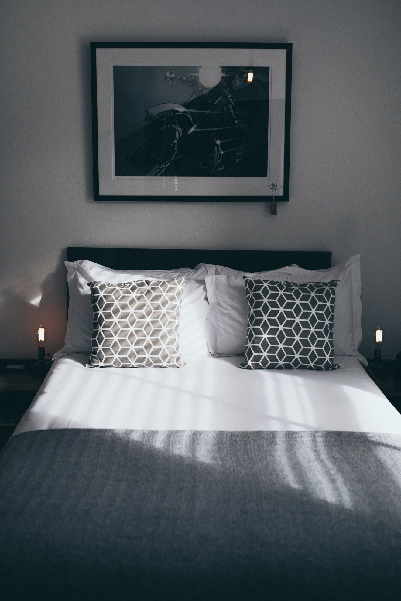 A neatly made bed with white linens and patterned pillows, a framed picture above it, and two bedside lamps.