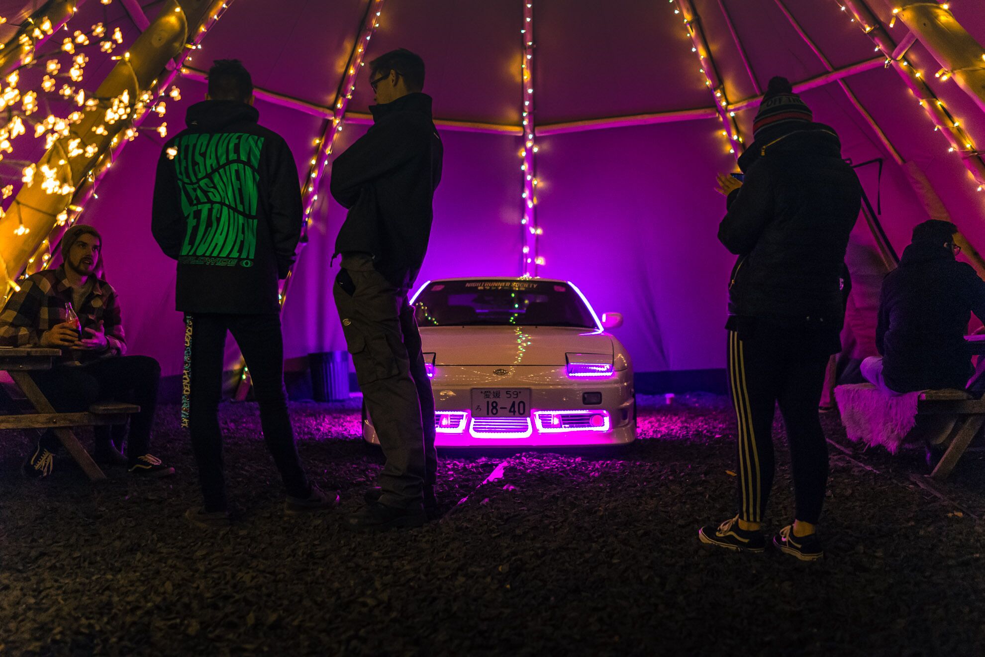 Una carpa con gente de pie alrededor de un coche con luces moradas.