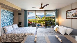 Een hotelkamer met een bed, een slaapbank, een balkon met uitzicht op strand en bergen, en een plafondventilator.
