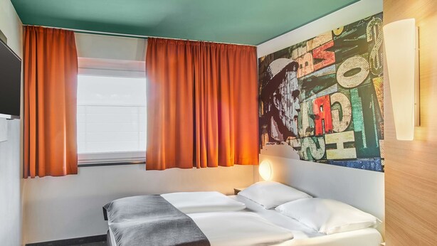 Une chambre d’hôtel avec un lit, un bureau, une fenêtre avec des rideaux et un mur orné d’une fresque colorée.