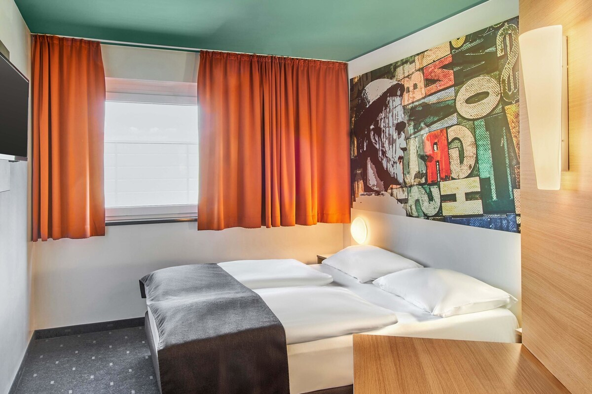 Une chambre d’hôtel avec un lit, un bureau, une fenêtre avec des rideaux et un mur orné d’une fresque colorée.