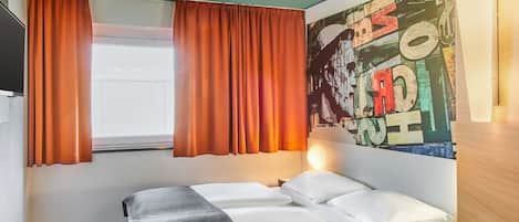 Una habitación de hotel con cama, escritorio, ventana con cortinas y un mural colorido en la pared.