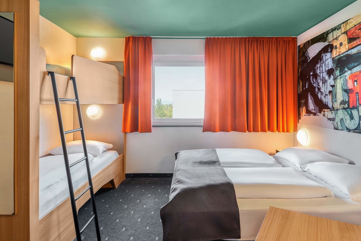 Une chambre d’hôtel compacte avec deux lits, un lit superposé, une échelle, une fenêtre avec des rideaux orange et un mur orné d’une fresque colorée.