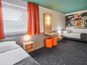 Une chambre d’hôtel moderne comprenant un lit, une table de chevet, une petite table d’appoint, une lampe fixée au mur, une grande fenêtre avec des rideaux et un tableau abstrait aux couleurs vives.