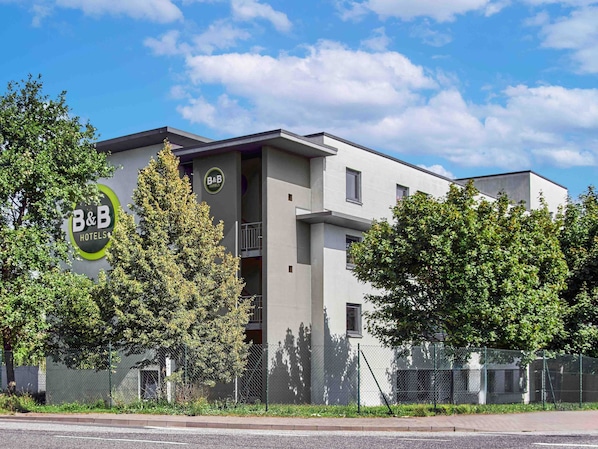 A modern B&B hotel with a grey facade, a balcony, and a fenced parking area.