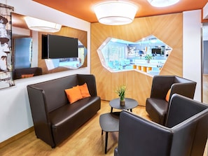 A modern lounge area with a black leather sofa, a round table, and two armchairs. A large mirror reflects the room, and a wooden accent wall with a geometric cutout is visible. - B&B Hotel Mainz-Hechtsheim (Mainz)