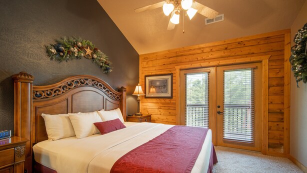 A bedroom with a wooden headboard, a large bed with white and red bedding, a nightstand, a lamp, a framed picture, and a window with blinds.