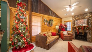 A cozy living room with a large Christmas tree, wooden walls, a stone fireplace, and a ceiling fan.