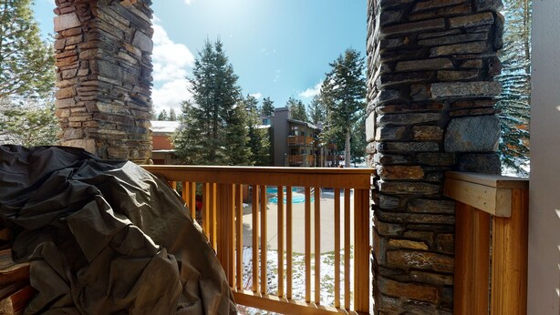 A wooden deck with a stone wall, a covered grill, and a view of a building with a pool. - Pet-Friendly Bright Corner Unit, Summer Pool, Tennis Courts, Hot Tub On-site (Mammoth Lakes)