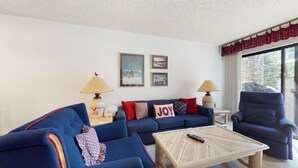 A living room with a blue sofa, a wooden coffee table, and a blue armchair. There are two lamps on side tables, a window with a view, and three framed pictures on the wall. - Pet-Friendly Bright Corner Unit, Summer Pool, Tennis Courts, Hot Tub On-site (Mammoth Lakes)
