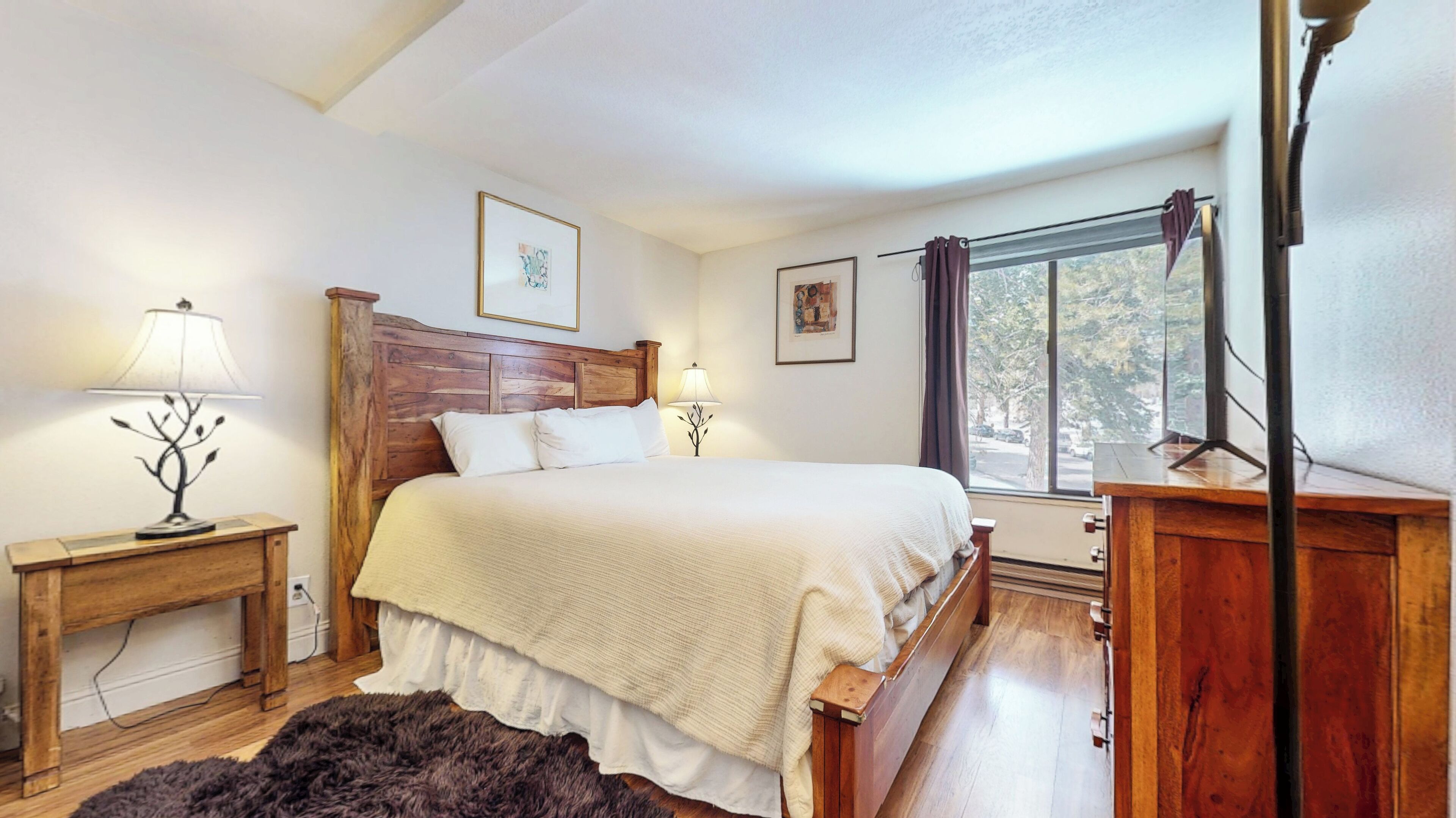 A bedroom with a wooden bed, two bedside tables with lamps, a window with curtains, and artwork on the walls.