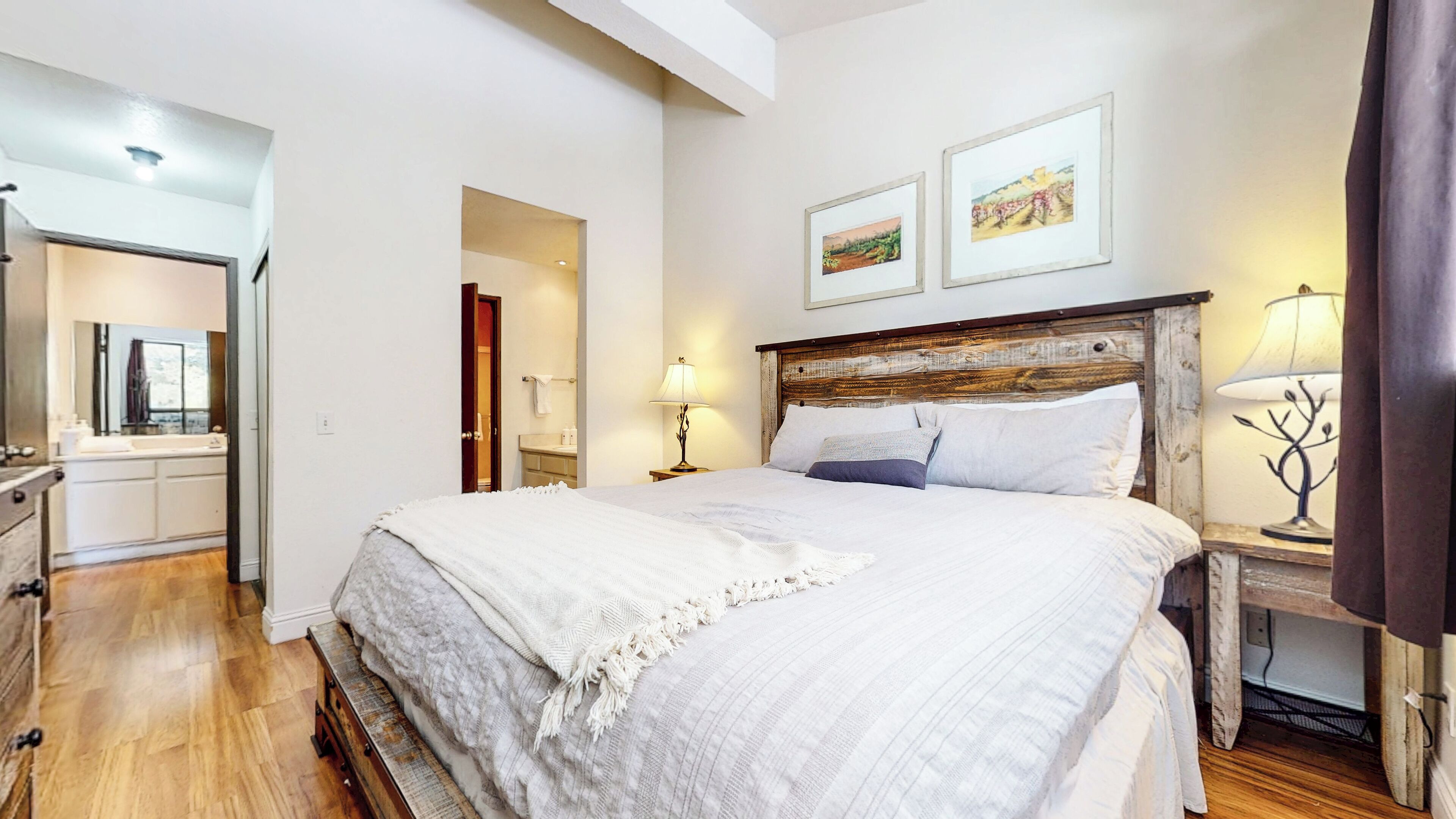 A bedroom with a wooden headboard, a large bed with white bedding, a nightstand with a lamp, and a bathroom visible through an open door.