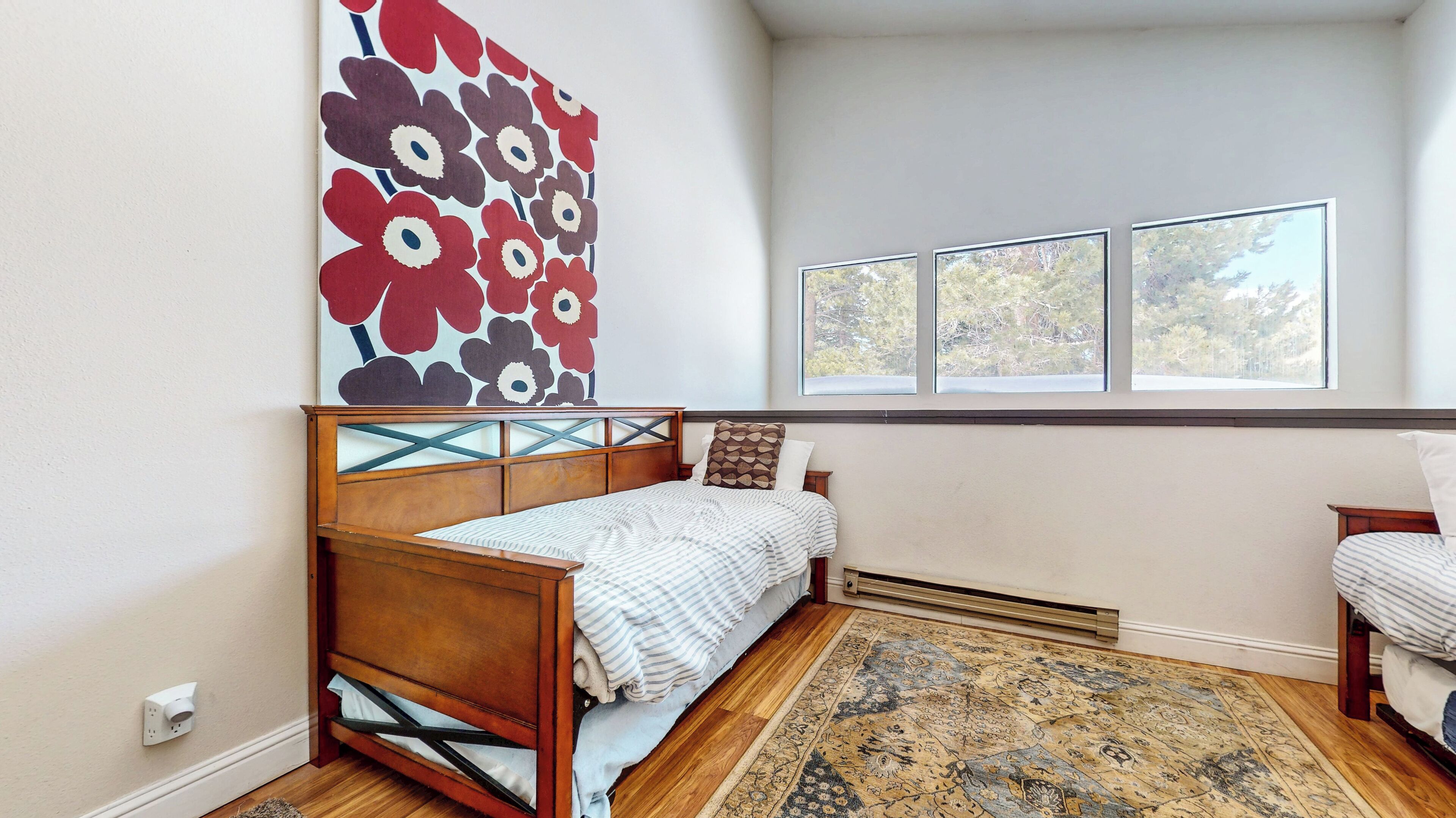A bedroom with a wooden bed, a patterned rug, a large floral artwork, and a window with a view of trees.