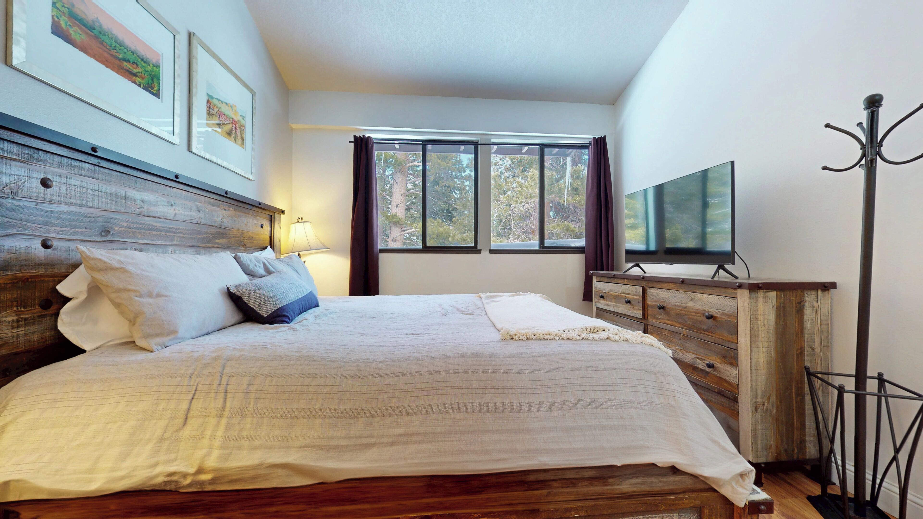 Un dormitorio con cabecera de madera, cama con ropa de cama blanca, televisor sobre un mueble de madera y una ventana con cortinas.