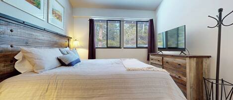 Un dormitorio con cabecera de madera, cama con ropa de cama blanca, televisor sobre un mueble de madera y una ventana con cortinas.