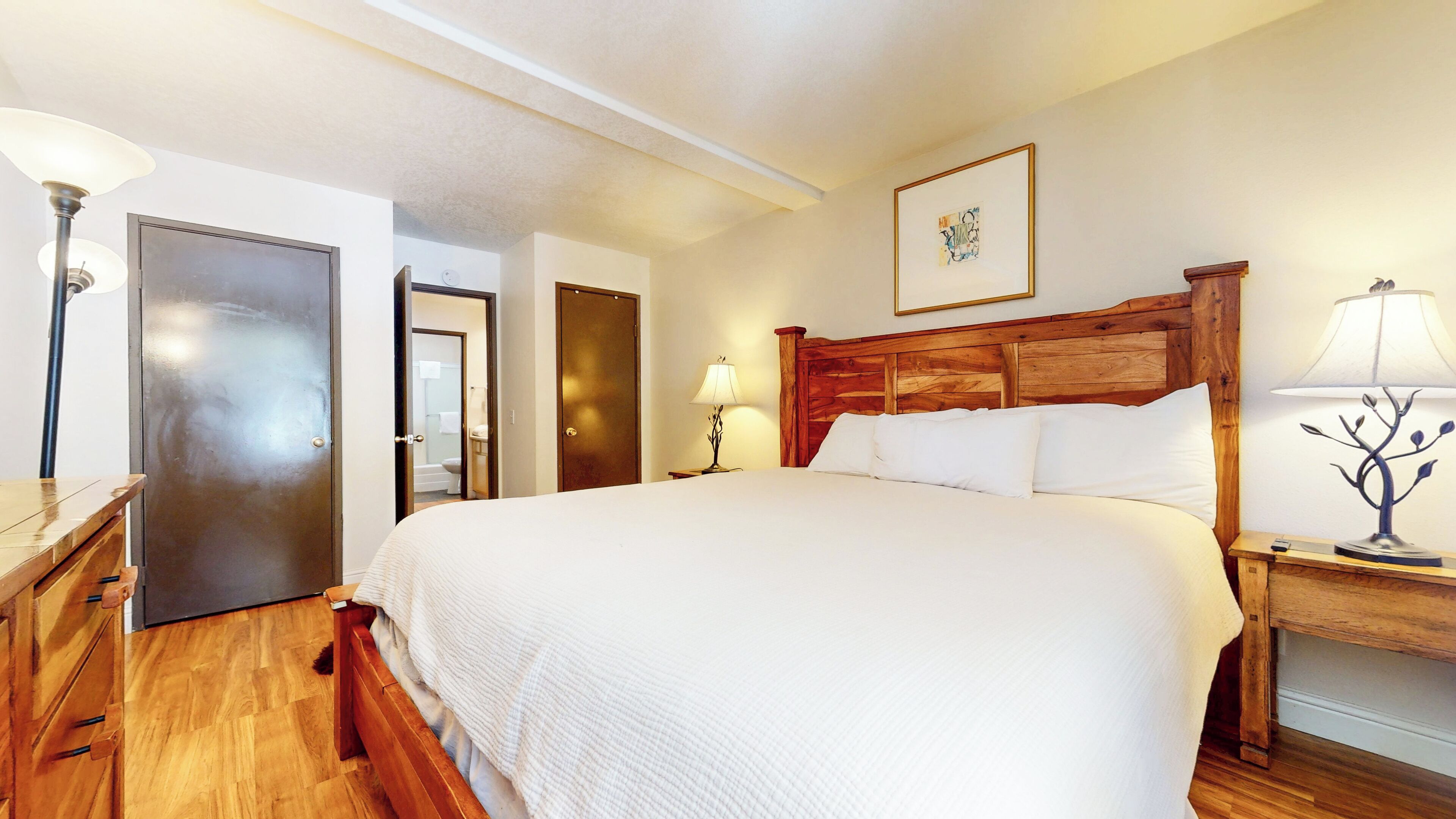 Een hotelkamer met een groot bed, een houten hoofdbord, een nachtkastje van hout met een lamp, een houten dressoir en een deur naar een badkamer.