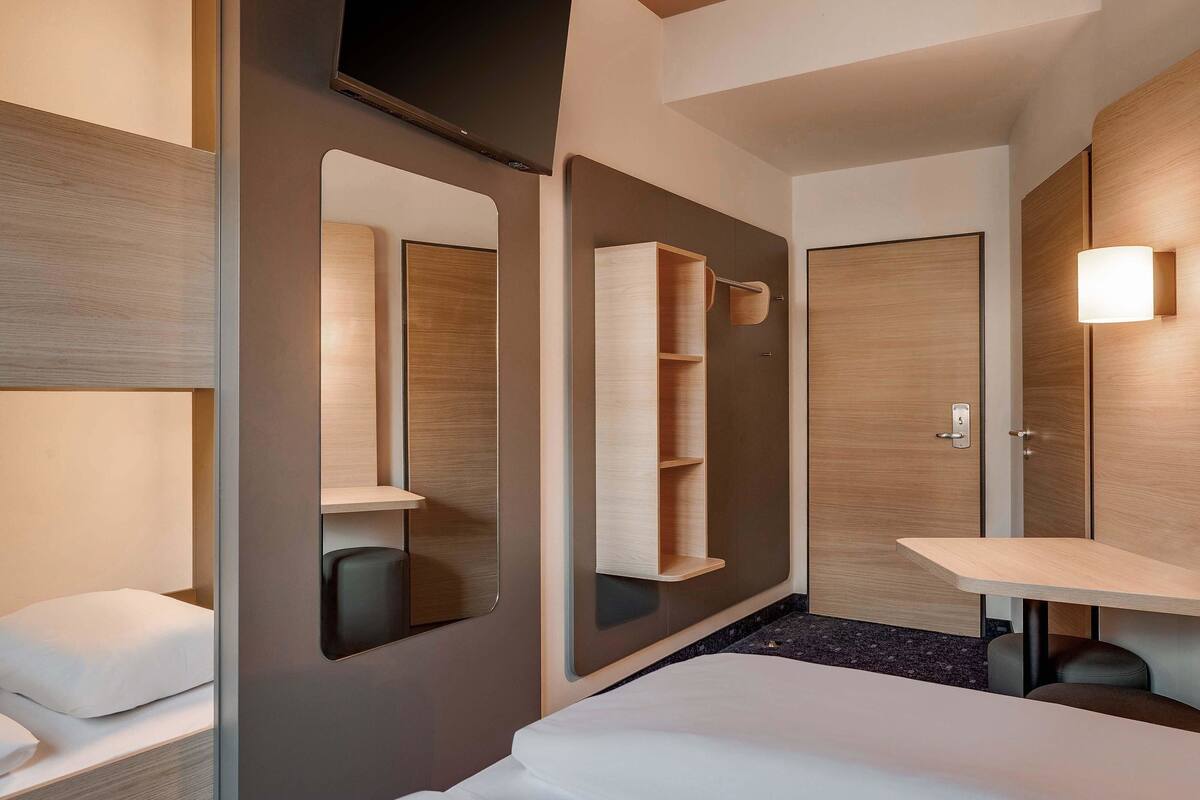 A modern hotel room with a bed, a large mirror, a bedside table, and a wardrobe.
