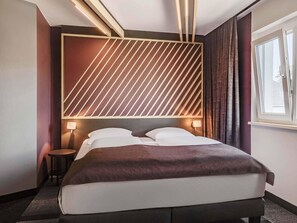 Habitación de hotel moderna con una cama grande, cabecera con líneas diagonales, lámparas de noche y una ventana con cortinas.